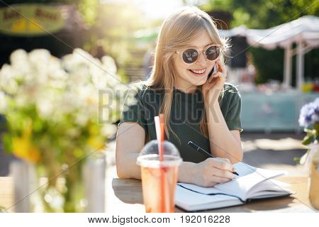 Portrait of a young business woman or student writing her plans in notepad talking on a smartphone smiling wearing glasses during a luch break in park on a sunny summer day. Education concept