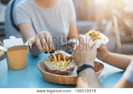 Close up of happy couple or friends coworkers having a burger and fries during lunchtime in a park on a sunny summer day. Junk food concept.
