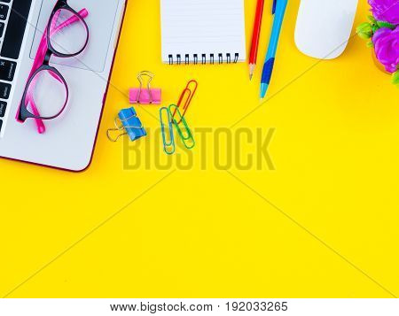 Flat lay top view office table desk frame. feminine desk workspace with office accessories including calculator mouse laptop glasses clips red pencil cutter note book blue pen and pink flower on yellow background.