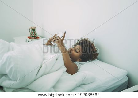 Beautiful curly young afro american girl has just woken up in the morning and checking e-mail and social networks using smartphone while laying sleepy on the white bed in her bedroom with red vase