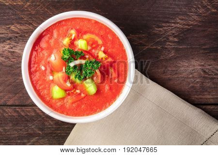 A bowl of gazpacho soup with slices of tomato, cucumber, and onion, with parsley leaves, shot from above on a dark rustic texture with a place for text