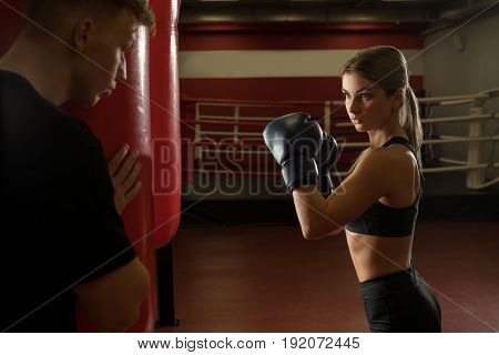 Sexy girl training in a gym in boxing gloves and her boyfriend hiding from her behind a punching bag