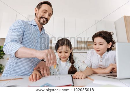 Learning through observing. Pleasant cheerful children sitting at the kitchen counter and learning how to use a compass while observing their father inscribing a circle