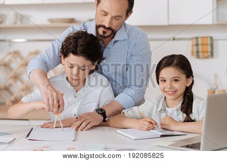 Help and care. Pleasant young man directing the hand of his son holding a pair of compasses while his cute cheerful daughter holding a pencil and looking at the camera with a broad smile