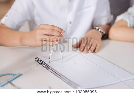 Moment of creation. The close up of delicate neat hands of a boy sitting at the table, holding a pair of compasses and inscribing circles