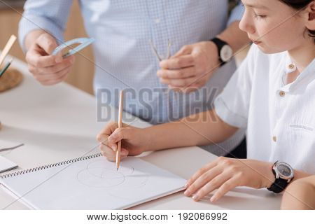 Focused on assignment. The close up of a cute little schoolboy drawing circles with a pencil while his father standing next to him and being ready to hand him a protractor and a pair of compasses