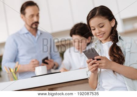 Happy communication. Charming little girl smiling and sending a text message to her friends while her father and brother being busy in the background