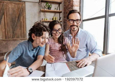 Hello there. Joyful single-parent family making a video call to their relatives while a cute little girl waving hello at the web camera