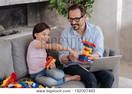 Nice idea. Handsome young father holding a house built with an erector set and watching an erector set assembling tutorial with his daughter while she pointing at the laptop with excitement