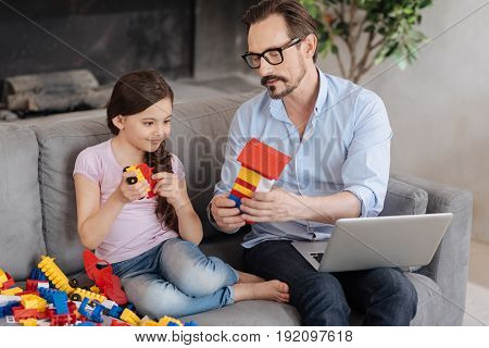 Look what I have. Pleasant young father sitting on the sofa and showing an erector set house to his beloved daughter while she holding a car model and looking at the house with smile