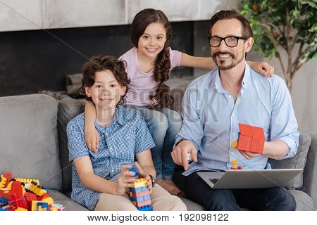 Sweet hugs. Gorgeous little girl sitting on the back of sofa and hugging her dear father and brother holding erector set pieces