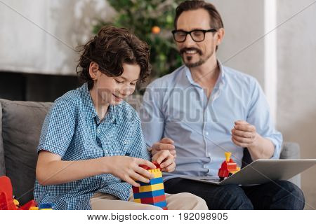Love new toy. Happy cheerful boy sitting on the sofa next to his father and assembling a tower with an erector set while his father talking to him
