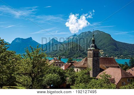 View of houses with belfry, in the village of Talloires, next to the Lake of Annecy. Mountains landscape on background, blue sky with clouds. France