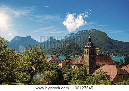 View of houses with belfry, in the village of Talloires, next to the Lake of Annecy. Mountains landscape on background, blue sky with clouds. France. Retouched photo