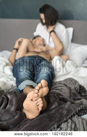 Cute girl in a white shirt hugs her boyfriend in blue jeans on the bed. He lies on her leg and looks at her face. Closeup low aperture photo. Vertical.