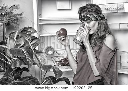 girl or woman hippie with cute face in green handkerchief or bandana on curly hair near fridge plant drink from glass and holds apple Black and white