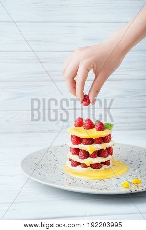 Delicious raspberry dessert with custard and orange caramel decorated by mint leaf on a beautiful plate and white wooden background. Tasty puff pastry standing on a table. Women's hand decorating it