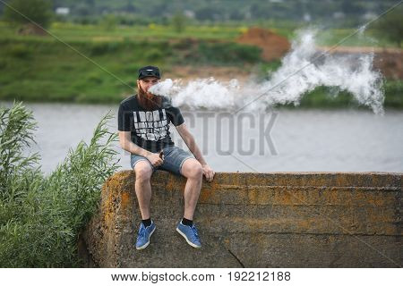 Vape Bearded Man In Real Life. Portrait Of Young Guy With Large Beard In A Cap And Sunglasses Having
