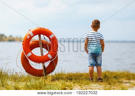 A little boy stands on the river bank near the rescue ring