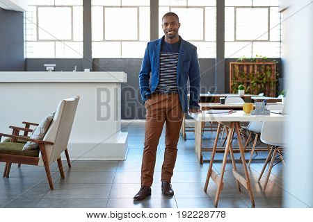 Portrait of a confident young African businessman in a blazer smiling while standing alone by a desk in a large modern office
