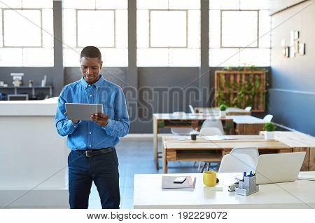 Focused young African businessman standing confidently in a large bright modern office working online with a digital tablet
