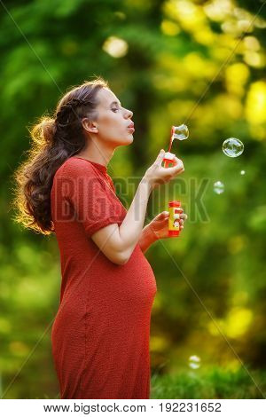 Pregnant woman in a red knitted dress blows soap bubbles standing on the edge of the forest at sunset