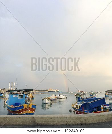 MARSAXLOKK, MALTA-June 1: traditional Maltese fishing boats with reflections and rainbow in he sunset sky background in maltese village MARSAXLOKK, MALTA in June 1,2017. Rainbow Marsaxlokk, Malta