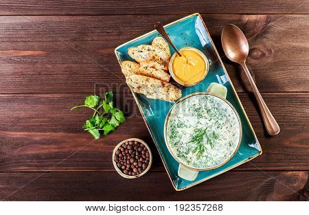 Cold soup or summer yoghurt with radish cucumber dill herbs and crackers on dark wooden background. Okroshka. Homemade food. Ingredients on table. Top view