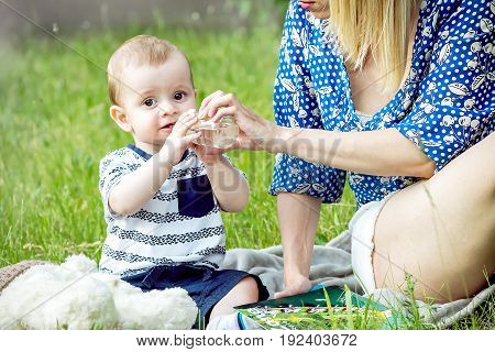 A pretty mather Giving water from a bottle with a pacifier to a baby boy sitting in a park on grass