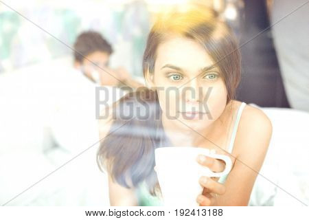 Thougtful woman having coffee at home with man lying in background
