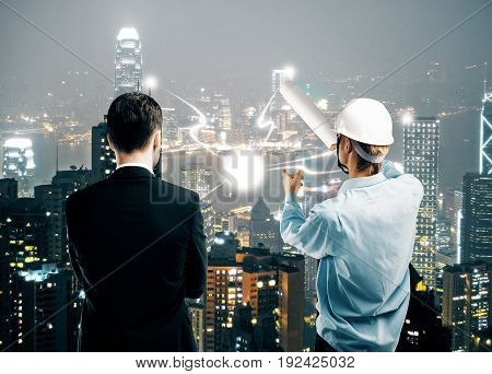 Back view of insdustrial worker explaining something to businessman on illuminated night city background. Architecture concept