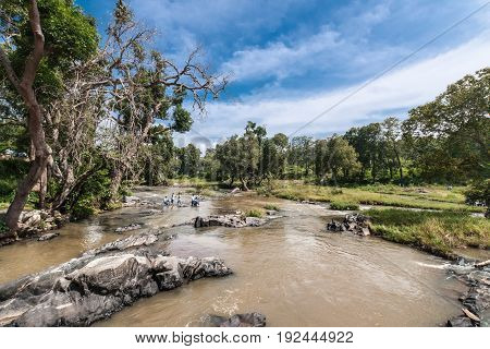 Tamil Nadu India - October 26 2013: Landscape shot of Moyar River in Masamigulli Forest. Brownish water flows over rocks through the green jungle under blue sky with white clouds. Far off students in photo.