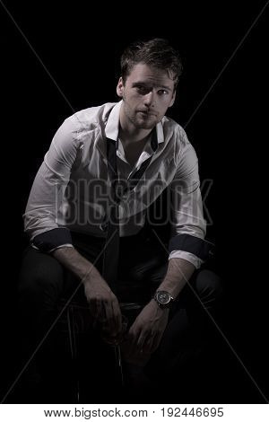 Portrait of young, attractive man in unbottoned, creased white shirt with upturned sleeves and loose tie sitting in the dark