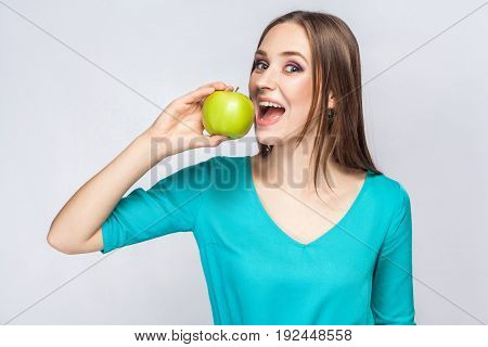 Young beautiful woman with freckles and green dress holding apple and eating looking at camera. studio shot isolated on light gray background.