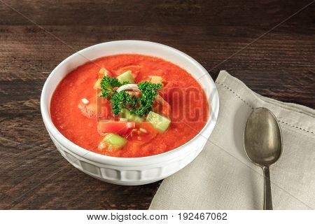 A bowl of gazpacho soup with slices of tomato, cucumber, and onion, with parsley leaves,  on a dark rustic texture with a place for text