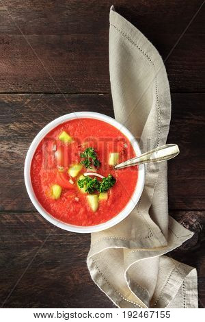 A bowl of gazpacho soup with slices of tomato, cucumber, and onion, with parsley leaves, shot from above on a dark rustic texture with a place for text