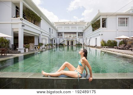 Portrait of relaxed young Caucasian woman wearing bikini sitting on pool edge at hotel and sunbathing