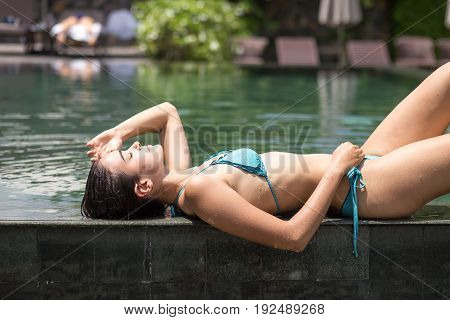 Portrait of serene young Caucasian girl wearing bikini lying at poolside and sunbathing. Young woman on vacation at summer resort