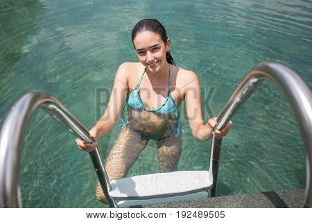Portrait of young Caucasian woman wearing bikini entering hotel swimming pool, looking at camera and smiling