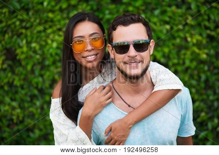 Closeup of smiling young Asian woman embracing Caucasian boyfriend from behind with green leaves wall in background. They are looking at camera. Front view.