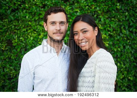 Closeup portrait of smiling young multi-ethnic couple looking at camera with green leaves wall in background
