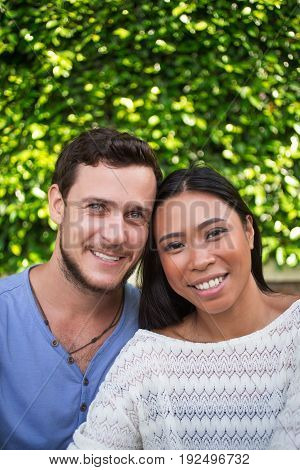 Closeup portrait of smiling young multi-ethnic couple looking at camera and sitting with blurred plants in background. Front view.