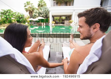 Closeup of young multi-ethnic couple looking at each other, holding hands and lying on chaise longues near swimming pool. They are back to camera.