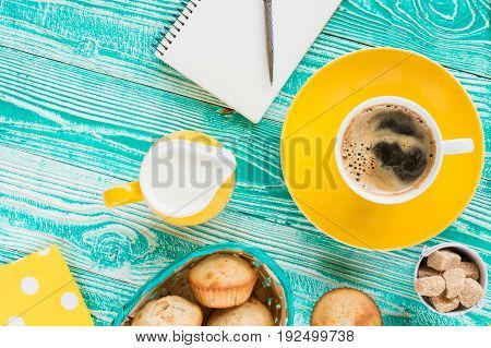 cup of coffee on yellow plate and yellow milk jug cane sugar notebook pen cakes teaspoon on turquoise colored wooden table with yellow napkin at polka dots top view