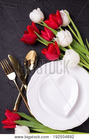 Festive table setting. White plate golden cutlery and white and red tulips flowers on black textured backgroud. Selective focus. Top view.