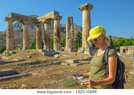 Tourism travel concept. Greece tourist female reads travel guide outdoors. Happy woman holding informative guide at Doric Temple of Apollo in Ancient Corinth, Peloponnese. Acrocorinth on background.