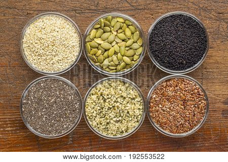healthy seed collection (chia, hemp hearts, brown flax, pumpkin, black cumin, sesame) - top view of small glass bowls against rustic wood
