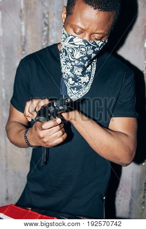 Black mercenary checks his weapon. Gangster man with gun in hand on dark gray background. Outlaw, ghetto, murderer, armed attack concept
