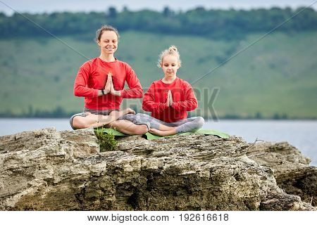 Sporty mother and daughter doing yoga on the rock near beautiful river. Beautiful mother and daughter dressed in the sportwear and barefoot. Horizontal photo with beauty landscape. Concept of the healthy and active lifestyle.