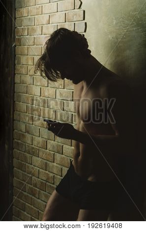 man or sexy caucasian macho with naked athletic torso in black trunks using smartphone or mobile phone on brick wall. Technology 4G 3G internet surfing. Fitness sport. Health diet